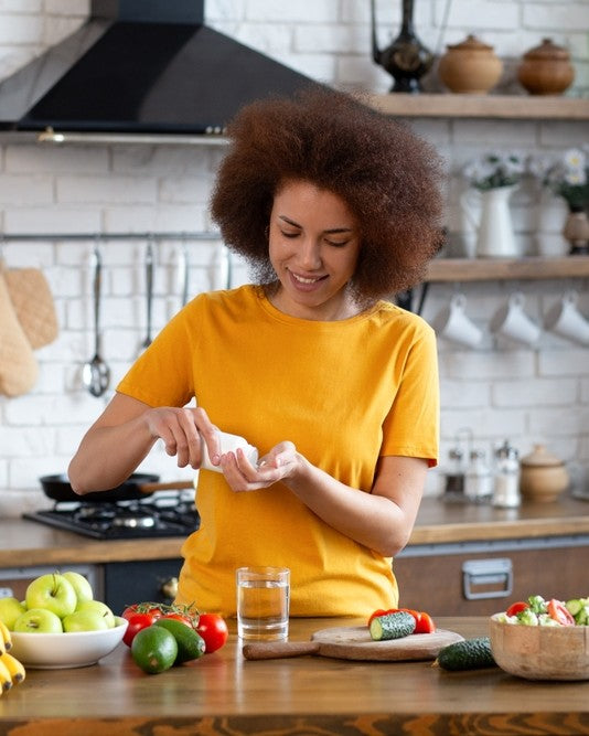 Woman in a yellow shirt preparing food in a kitchen with various fruits and vegetables on the counter.